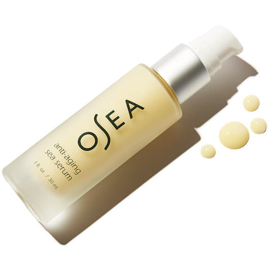 A bottle of osea anti-aging sea serum with a pump dispenser lying on a white surface, accompanied by small drops of the serum.