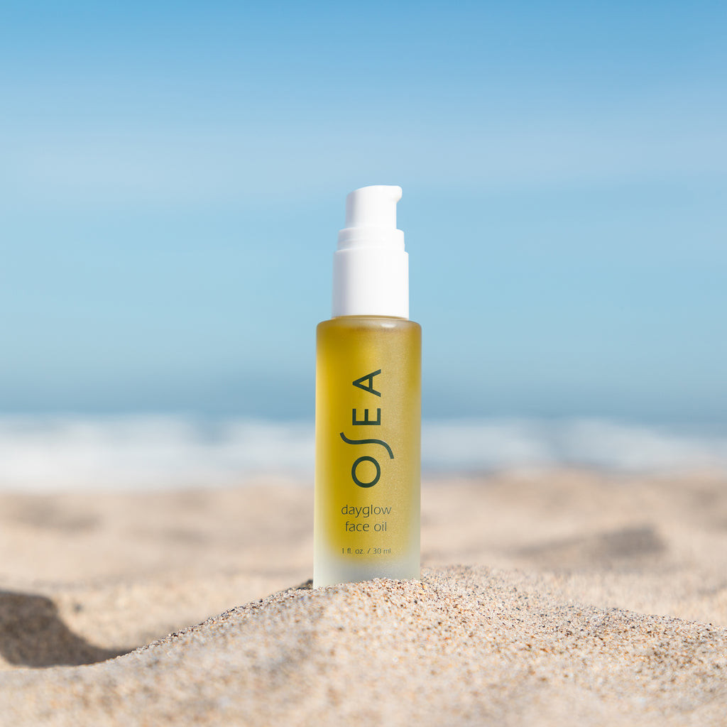 Bottle of facial oil on a sandy beach with a clear blue sky in the background.