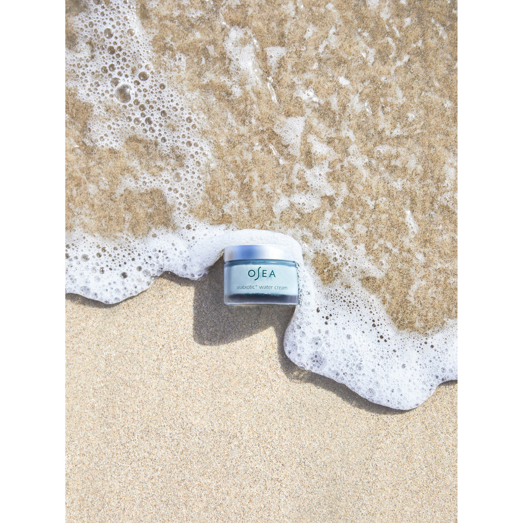 A skincare product jar rests on sandy beach as gentle waves approach it.