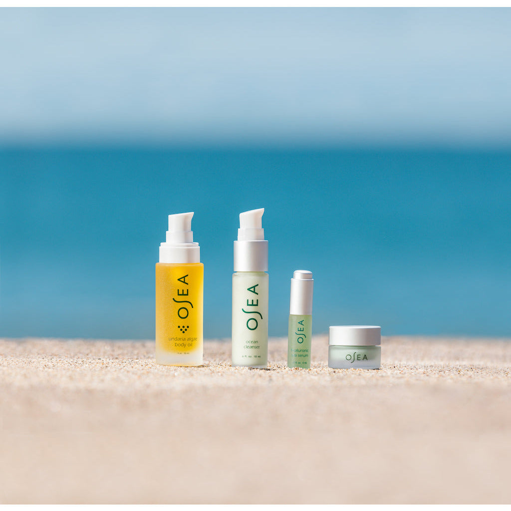 Four skincare products arranged on a sandy beach with a clear blue sky and tranquil sea in the background.