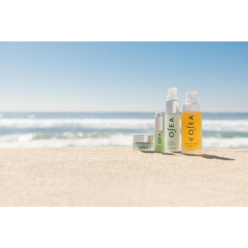 A collection of skincare products arranged on a sandy beach with the ocean in the background.