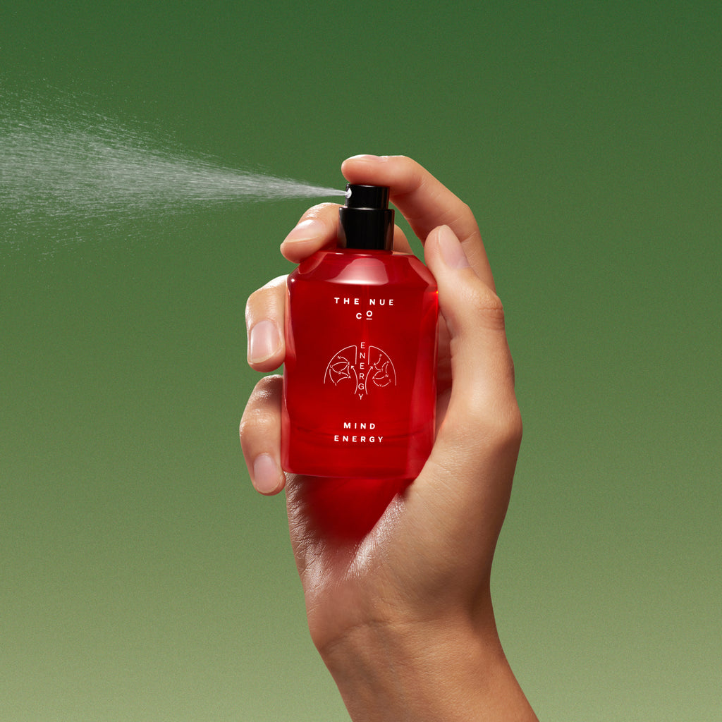 A hand holding a red bottle with a mist spraying out against a green background.