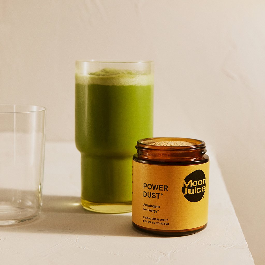 A jar of 'power dust' supplement next to a glass of green juice on a neutral background.