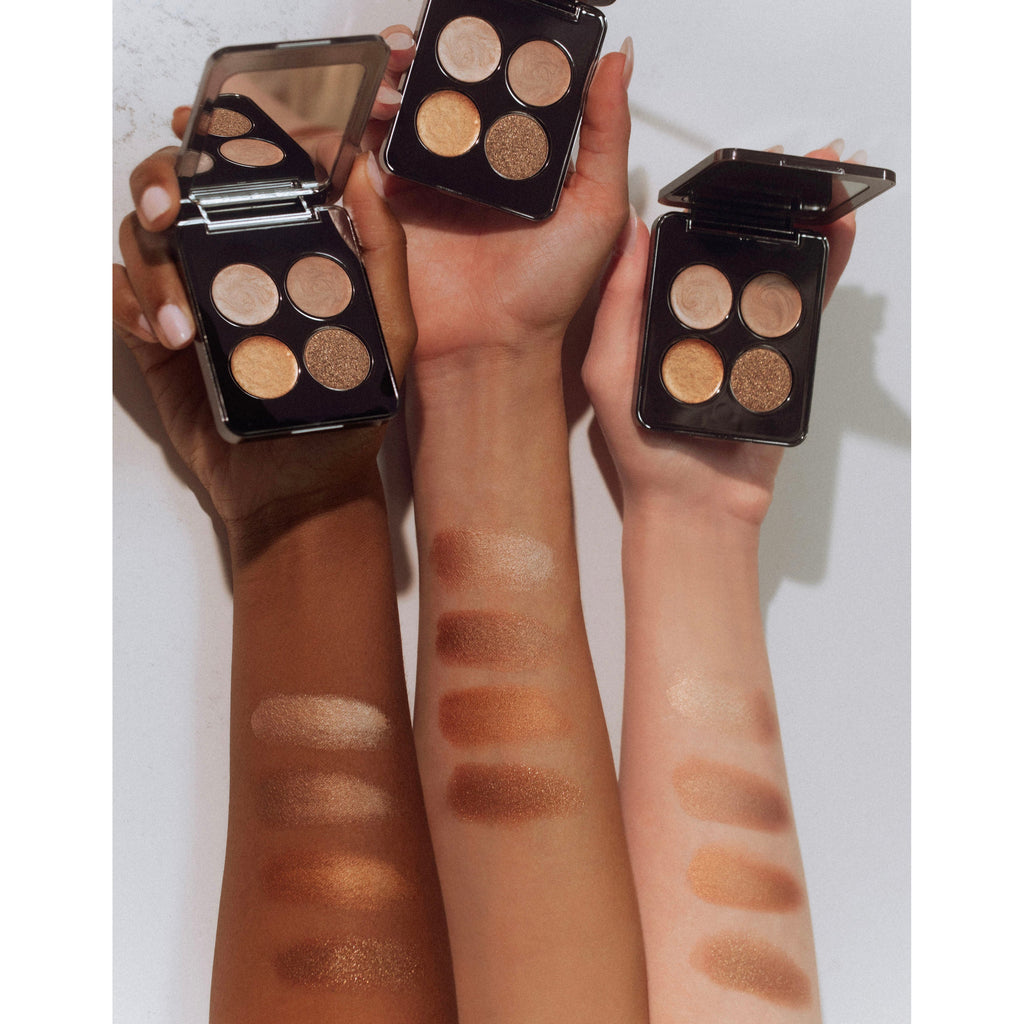 Three arms with different skin tones showing swatches of eyeshadow shades from palettes held in hands.