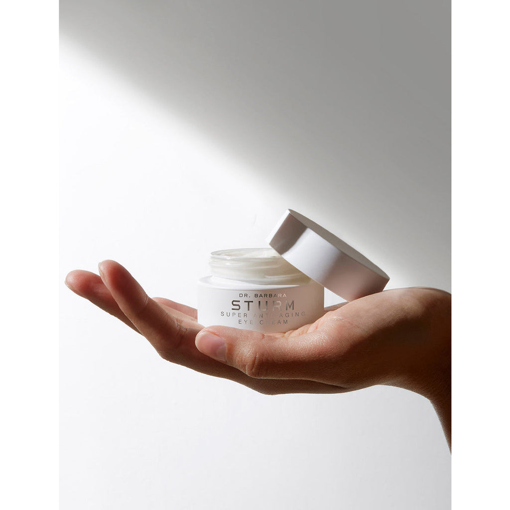 A hand holding a jar of dr. barbara sturm eye cream with the lid off, against a white background with a shadow.