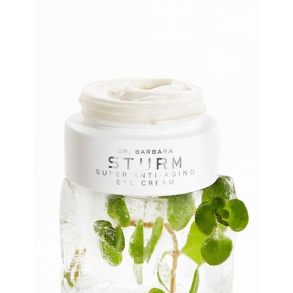 Open jar of eye cream on ice with green leaves.
