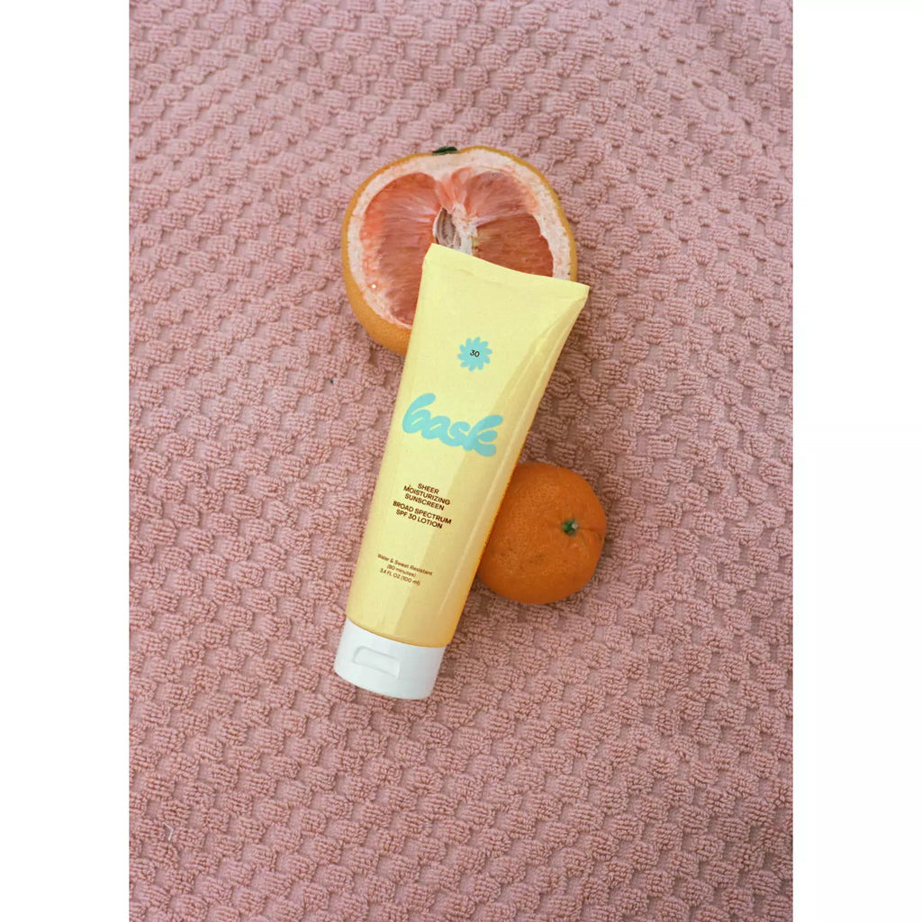 A tube of sunscreen with a floral logo beside a sliced grapefruit and a whole tangerine on a textured pink background.