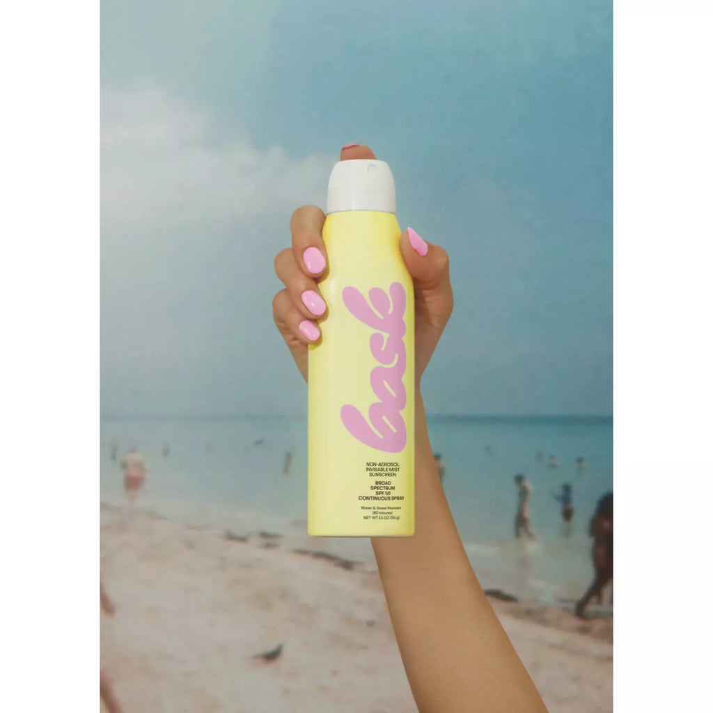 A person holding up a yellow sunscreen spray bottle against a beach backdrop.