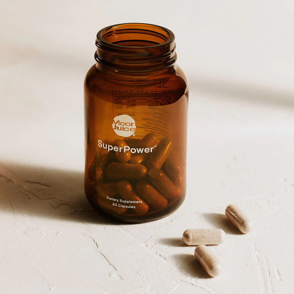 A jar of dietary supplement capsules labeled "superpower" by moon juice on a light background, with three capsules lying outside the jar.