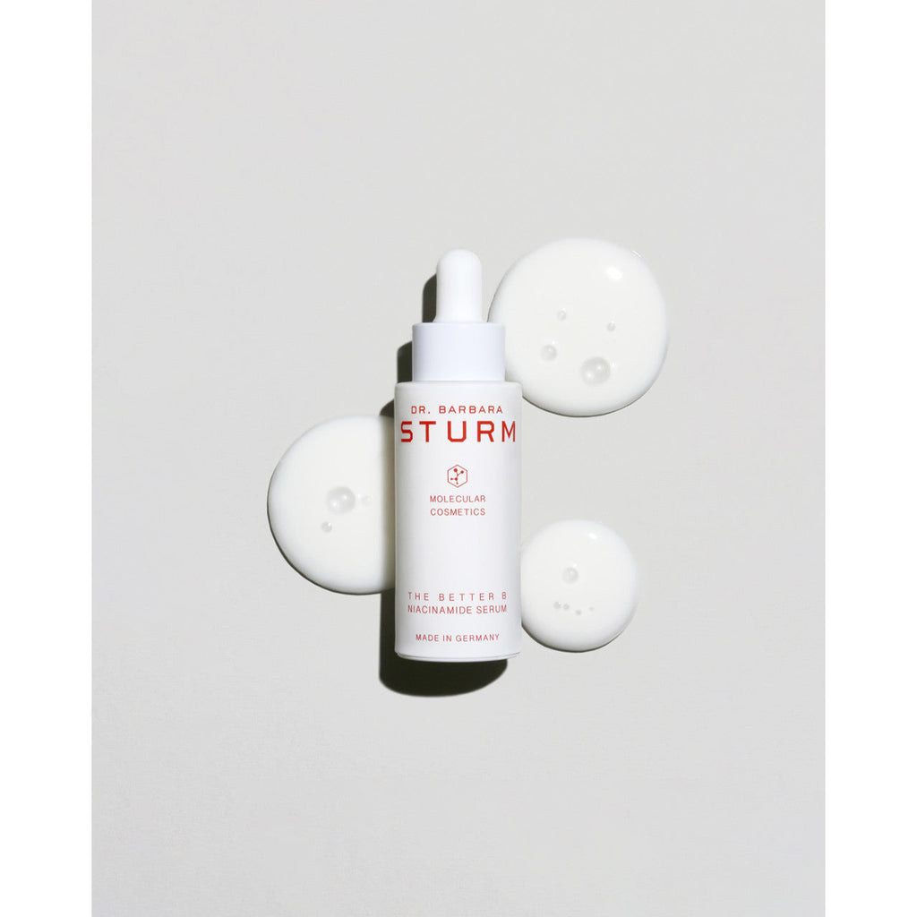 A bottle of dr. barbara sturm skincare serum with droplets around it on a light background.