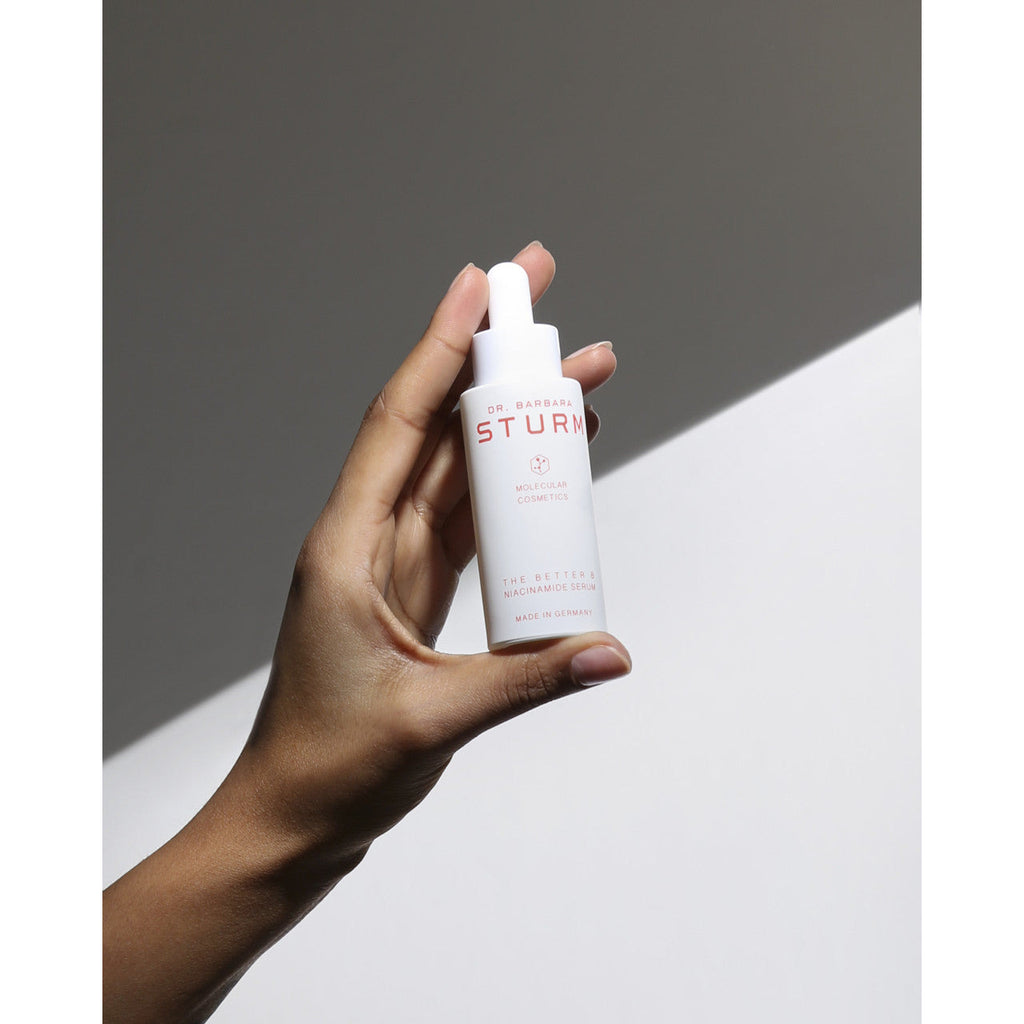 A hand holding a bottle of dr. barbara sturm skincare product against a two-tone background.