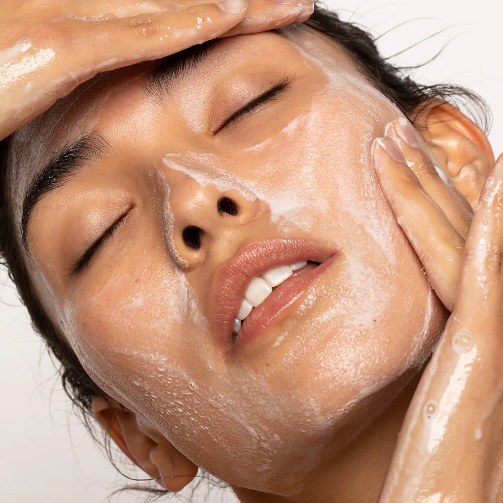 A person applying facial cleanser with their eyes closed and water droplets on their skin.