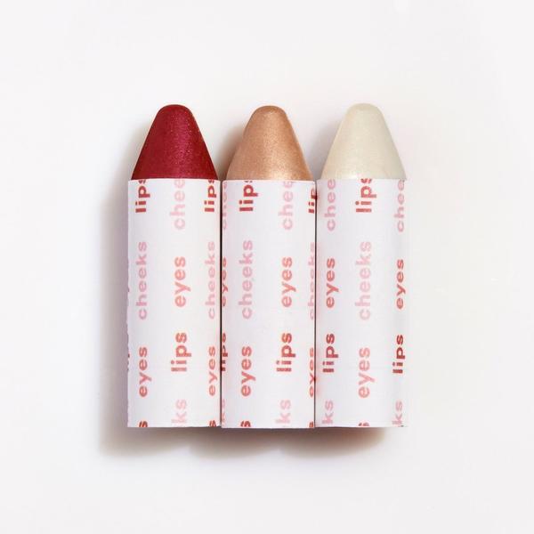 Three crayon-shaped cosmetic sticks with shades of red, bronze, and white, labeled for lips, cheeks, and eyes.