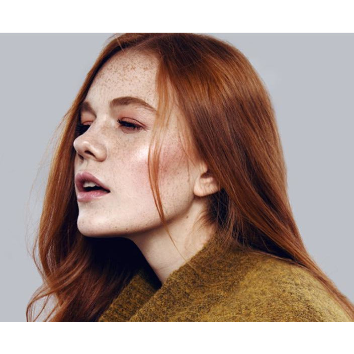 A woman with red hair and freckles wearing a mustard-colored garment looks to the side with a neutral expression.