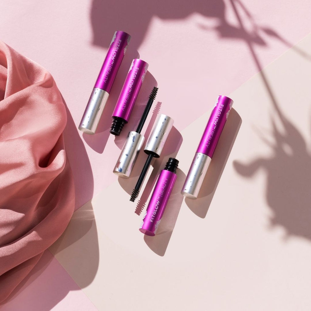 A collection of pink mascara tubes artfully arranged on a pink surface with soft shadows and a draped fabric corner.