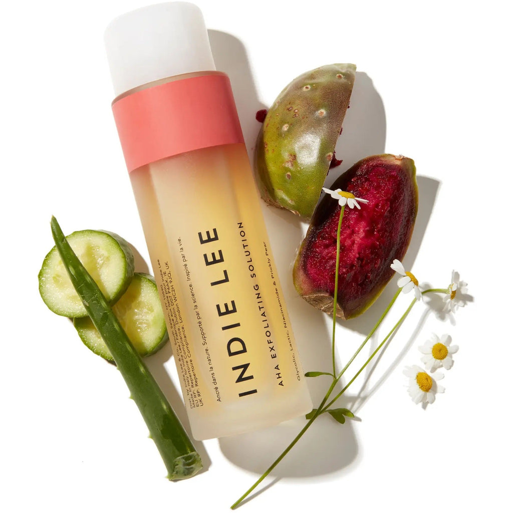 Bottle of indie lee skincare product with natural ingredients around it, including slices of cucumber, a halved prickly pear, and small white flowers.