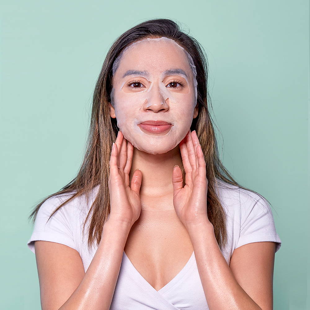 Woman applying a facial sheet mask and touching her face gently against a green background.