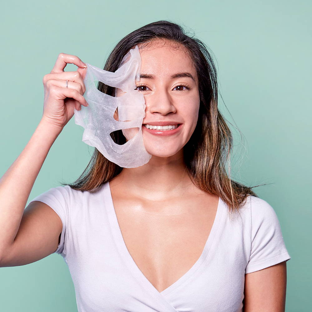 A woman holding a facial mask next to her face and smiling at the camera.