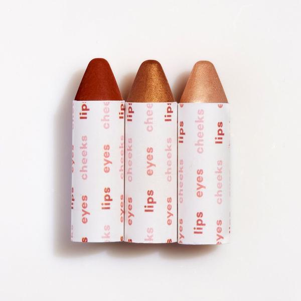 Three crayon tips in shades of red and brown, partially wrapped in paper labeled for lips, cheeks, and eyes, suggesting they are multi-purpose cosmetic crayons.