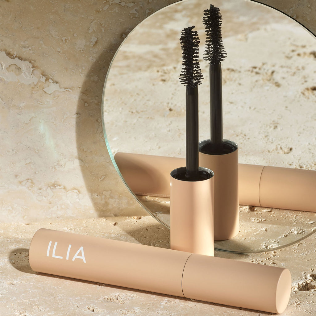 A mascara tube and wand from ilia beauty brand reflected in a circular mirror on a textured surface.
