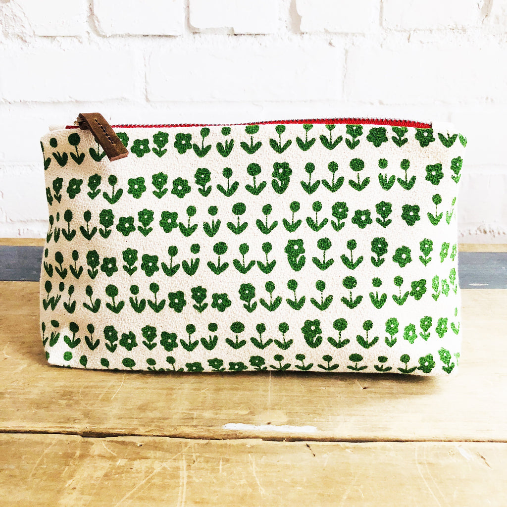 A fabric pouch with a green floral pattern on a wooden surface against a white brick wall.
