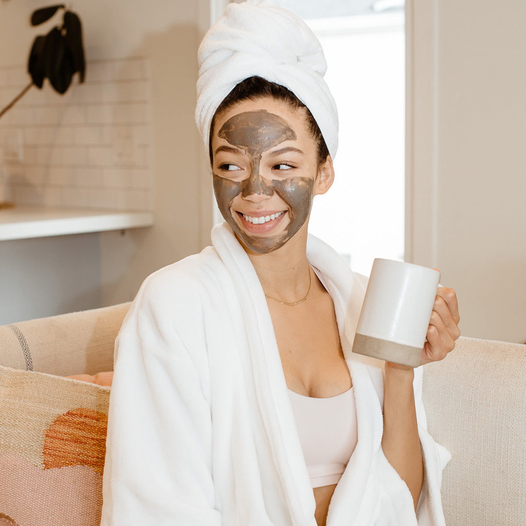 Woman in a robe and facial mask enjoying a cup of coffee on the couch.