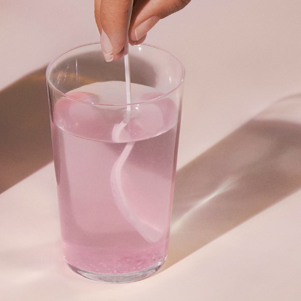 A person stirring a pink beverage in a clear glass with ice cubes.