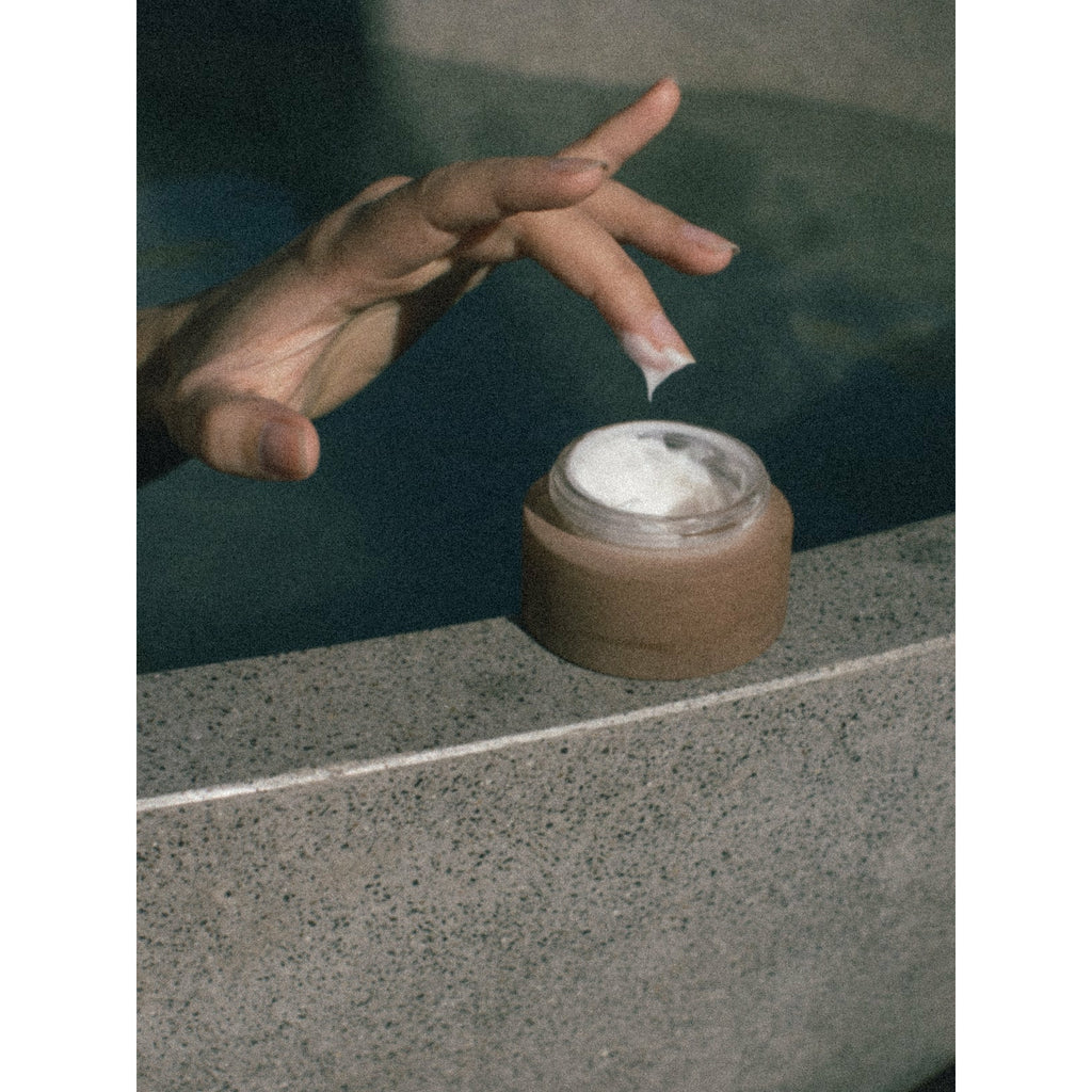 A hand reaching for a cosmetic cream in an open container positioned on a ledge.