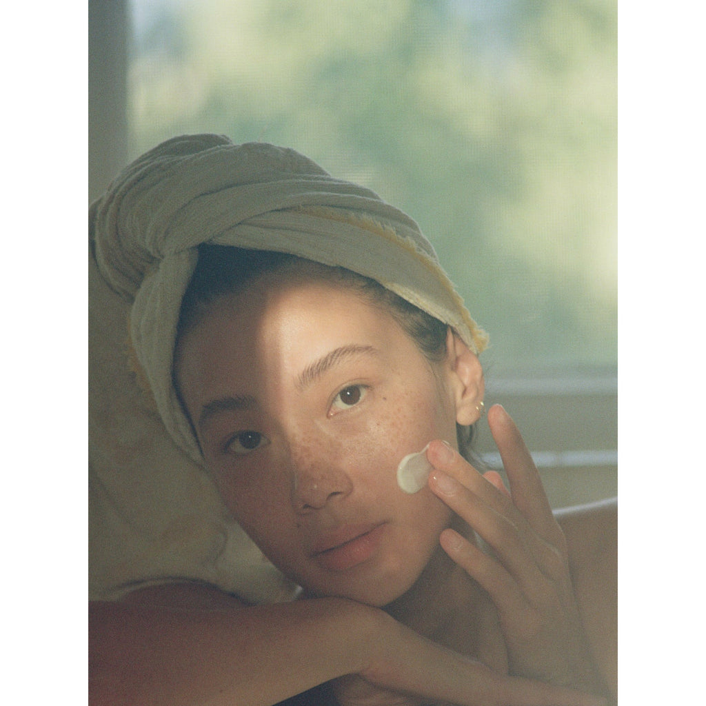 A person with a towel wrapped around their hair applies cream to their face in a sunlit room.