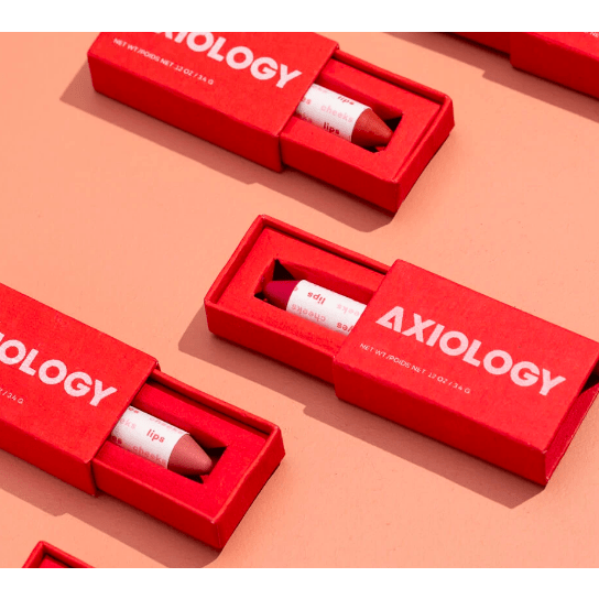 Three red lipstick boxes with the brand name "axiology" on a pink background, with one box open revealing a lipstick tube inside.