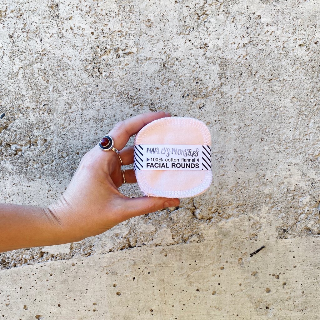 A hand holding a pack of pink reusable cotton facial rounds against a concrete background.