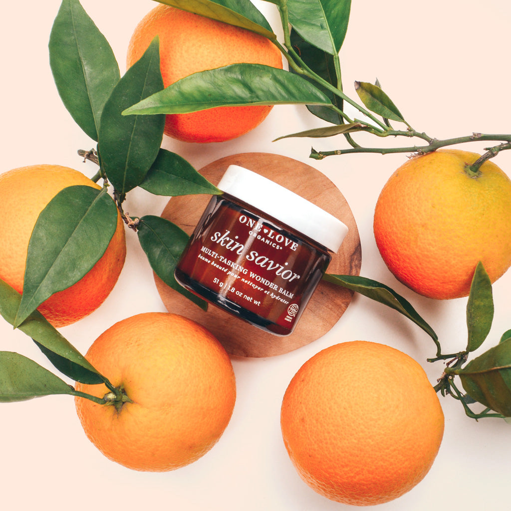 Jar of skin care product labeled "skin savior" surrounded by fresh oranges and green leaves on a wooden surface against a pale background.
