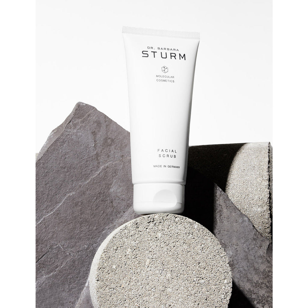 A tube of dr. barbara sturm facial scrub placed on a textured surface among geometric stone shapes, highlighted by contrasting shadows.