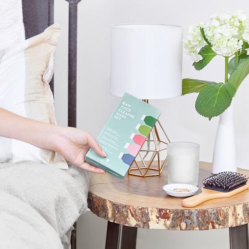 Person holding a skincare product beside a lamp and vase on a wooden side table.