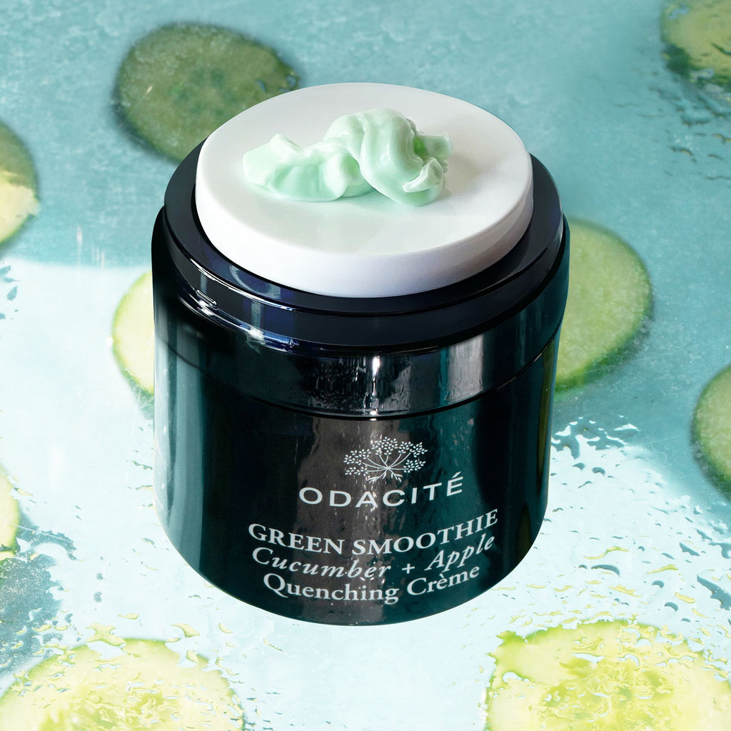A jar of odacite green smoothie quenching crÃ¨me with a cream smear on its lid, surrounded by slices of cucumber and splashes of water.