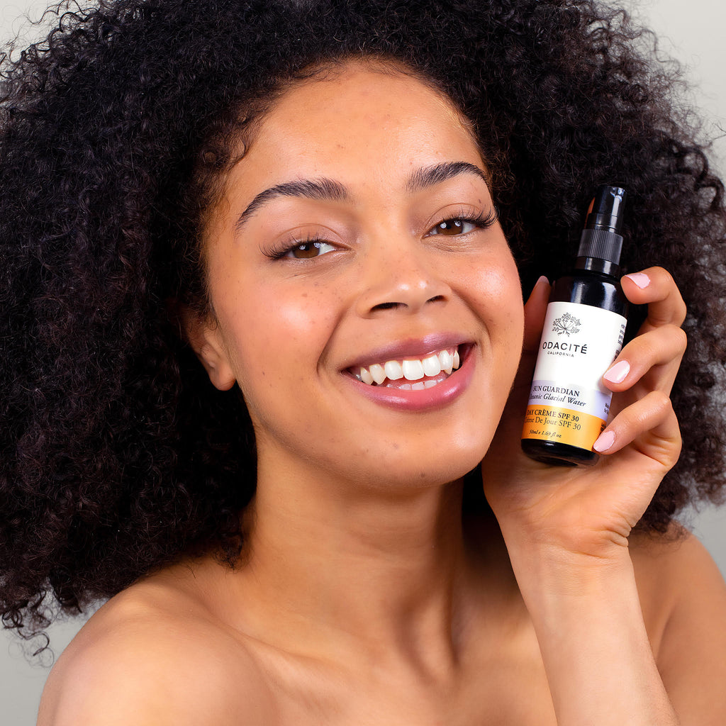 Woman with curly hair smiling and holding a skincare product bottle.