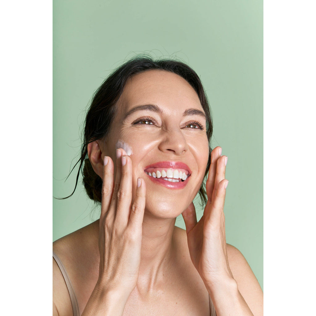 A woman smiling while applying cream to her face against a green background.