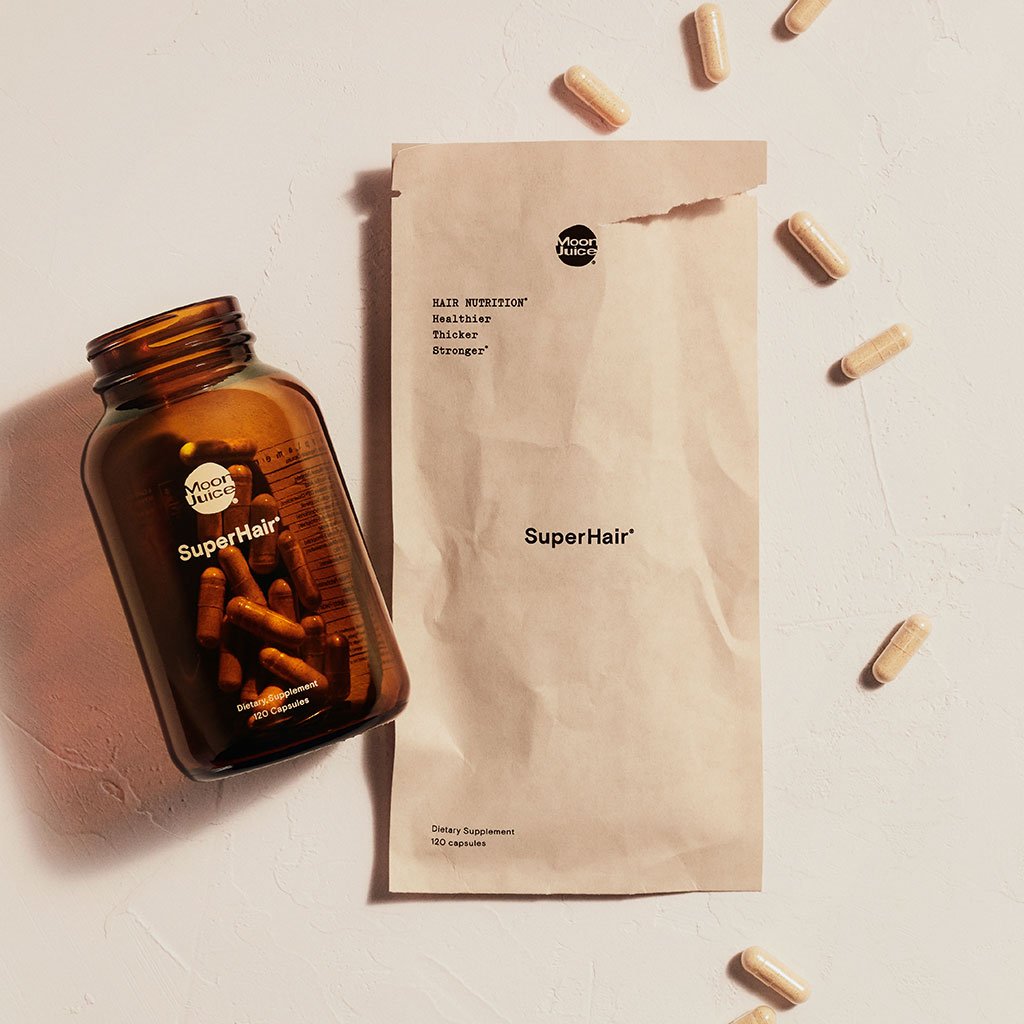 A bottle of "superhair" dietary supplements next to a paper bag and scattered capsules on a textured surface.