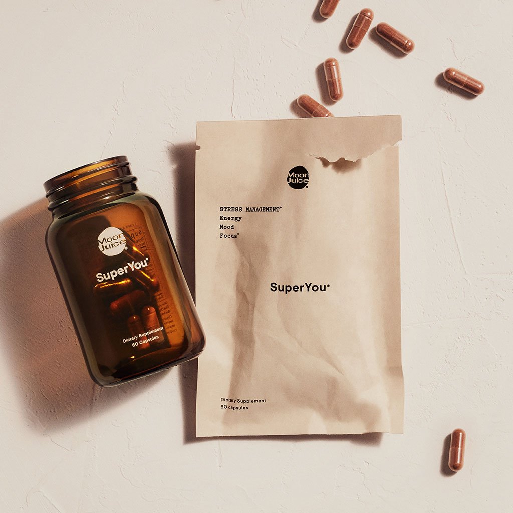 A bottle of "superyou" dietary supplements by moon juice alongside a paper bag and scattered capsules on a textured surface.