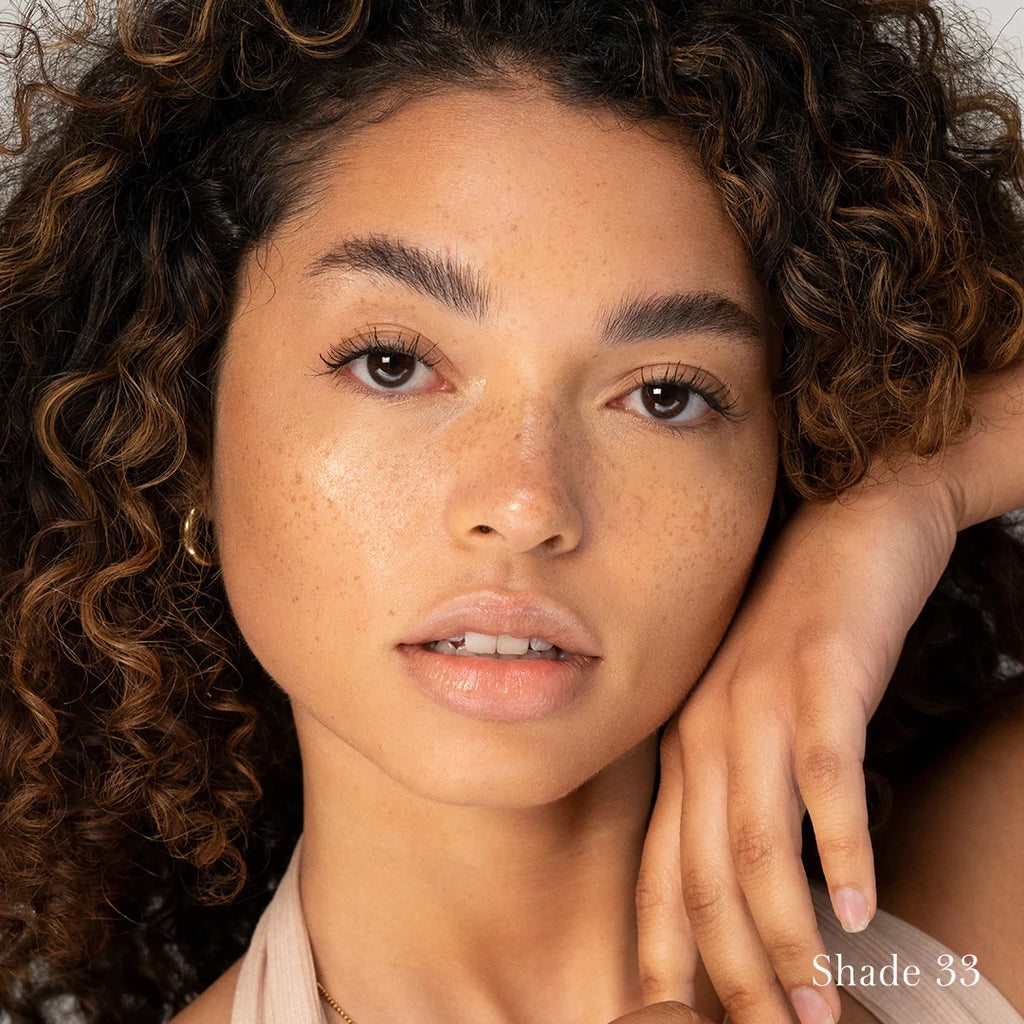 A woman with curly hair showcasing a natural makeup look, highlighting her freckles and featuring "shade 33.
