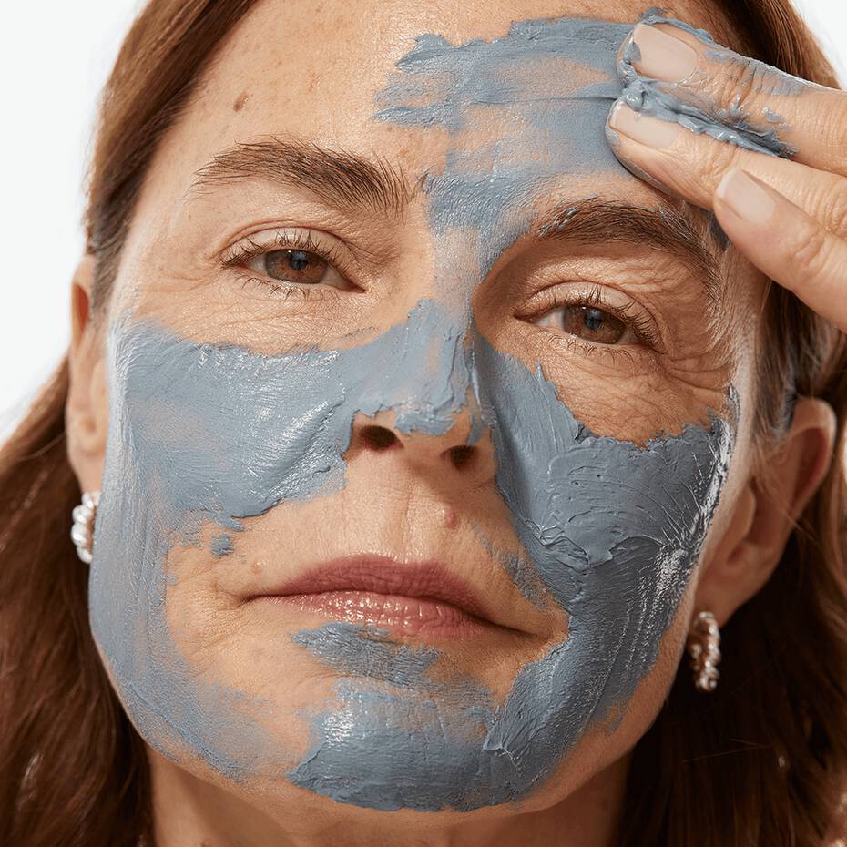 A woman applies a gray clay mask to her face.