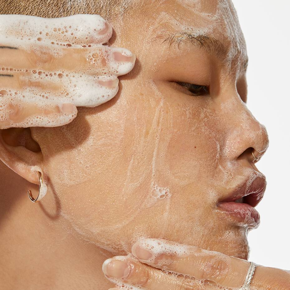 A person cleansing their face with a foaming cleanser, focusing on the cheek and temple area.