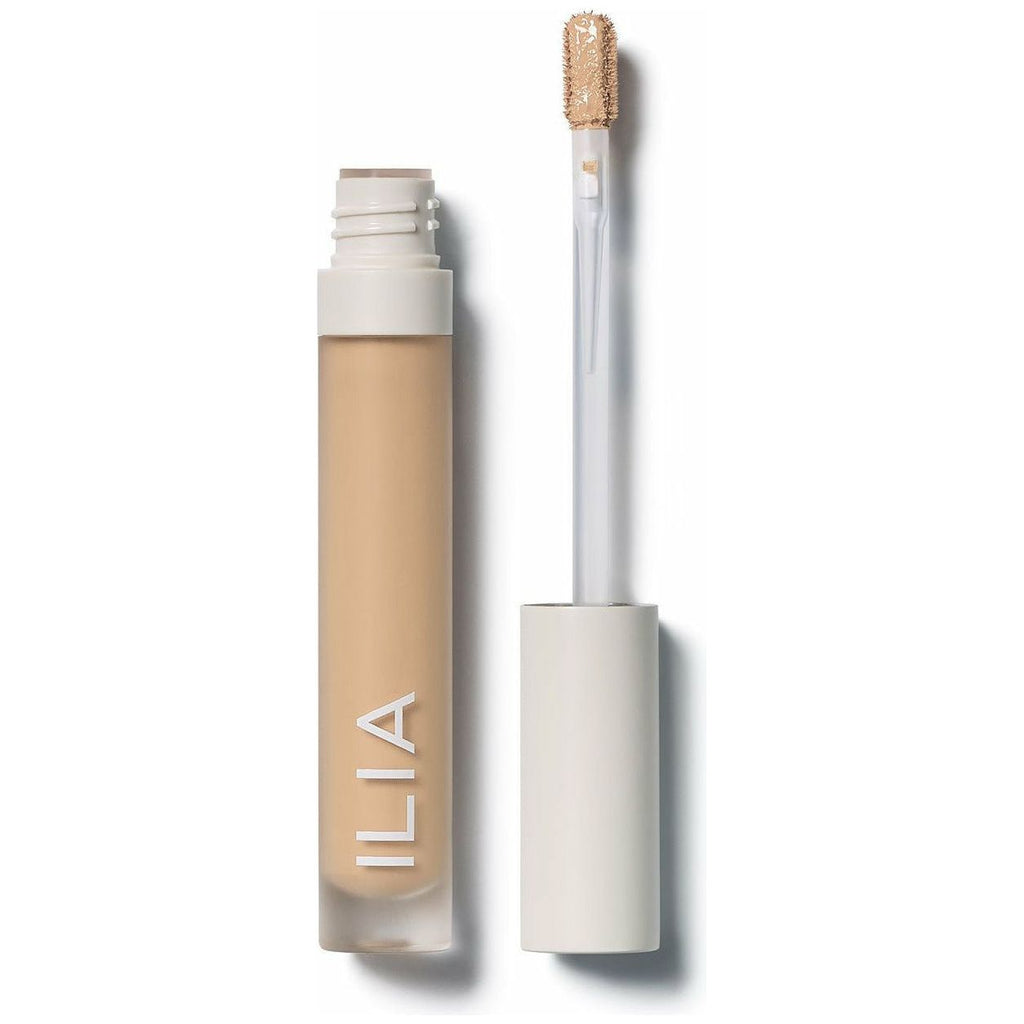 A bottle of ilia brand concealer with its cap and brush applicator removed and positioned to the side.